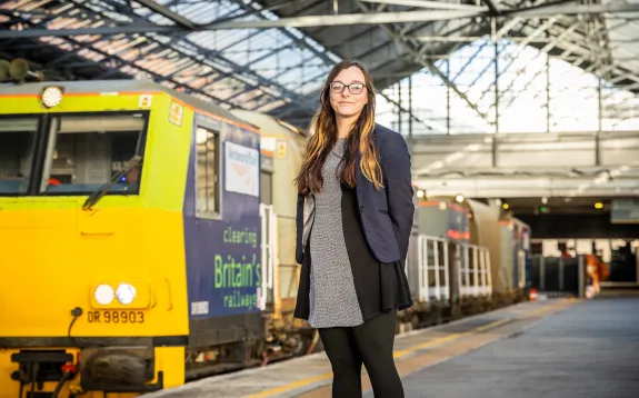Image shows a woman standing on a train platform in front of the leaf buster train. She is wearing a black and grey dress, black tights a navy blue blazer and glasses.
