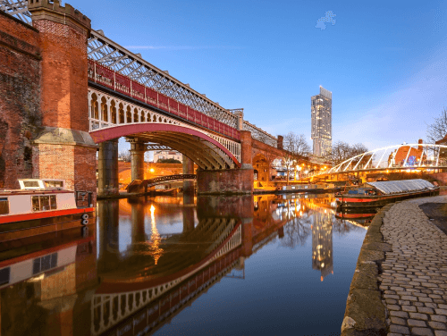 Evening shot of the River Medlock with a historic red bridge and canal boats sailing along the river